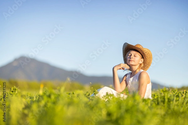 Fototapeta Young joyful girl on a blooming meadow with a hat on her head at sunset. Spring joyful mood child in the grass
