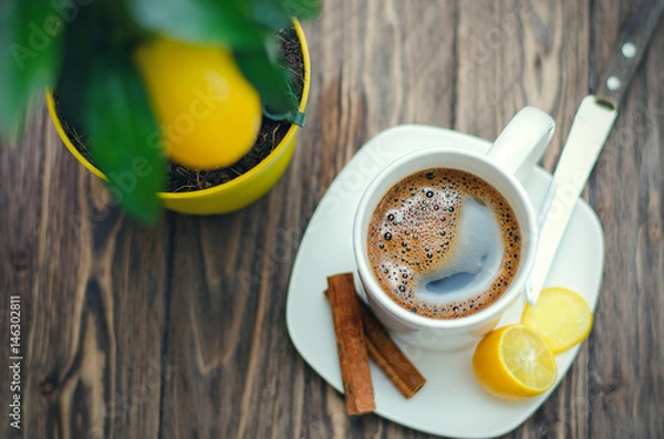 Fototapeta Coffee in white cup on wooden table opposite a defocused background with lemon tree.