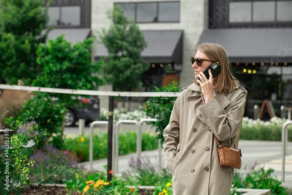 Obraz Woman in trench coat talking on phone in urban garden setting. Businesswoman 
