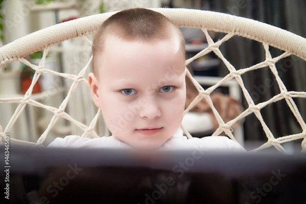 Fototapeta Child focused on a screen while sitting in a macrame chair at home during the afternoon
