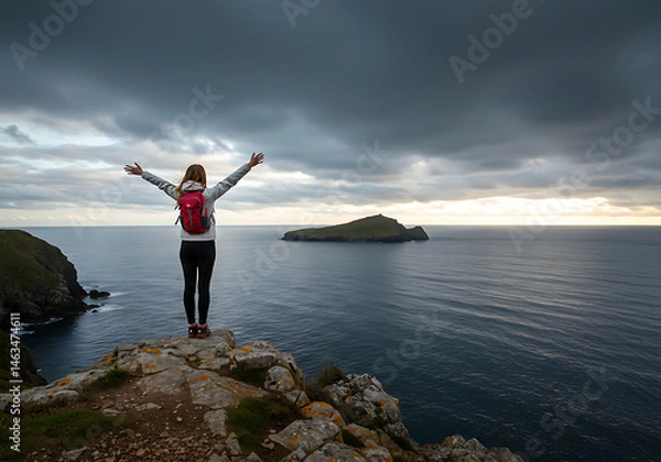 Fototapeta A woman with a backpack stands on a rocky cliff edge, arms outstretched, admiring a dramatic coastal view with the sea and a distant island under a cloudy sky