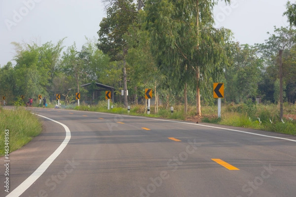 Fototapeta view of a curved asphalt road winding through a rural landscape in Thailand. Several yellow and black curve warning signs are visible along the roadside