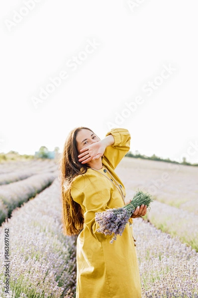 Fototapeta woman in yellow dress stands in lavender field, gently holding bouquet of lavender flowers. She appears serene and contemplative, enjoying beauty and fragrance of blossoms around her.