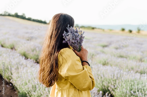 Fototapeta woman in yellow dress stands in lavender field, gently holding bouquet of lavender flowers. She appears serene and contemplative, enjoying beauty and fragrance of blossoms around her.