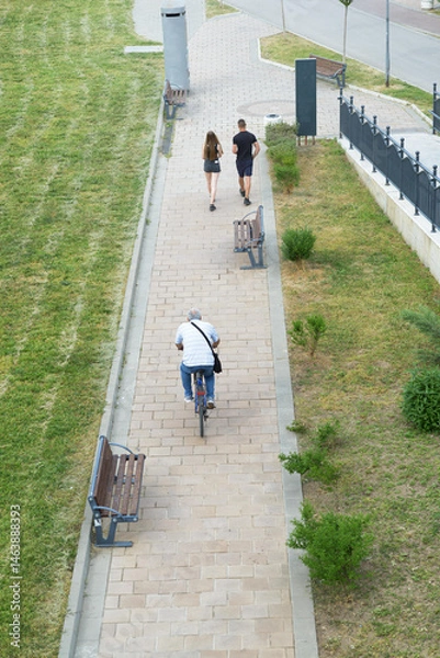 Fototapeta A senior man cycles while a young couple walks along a paved urban park pathway bordered by benches and grass under bright daylight.