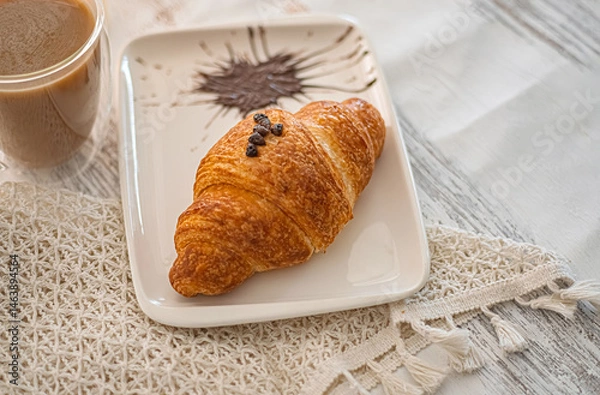 Obraz Freshly baked croissant with coffee. A croissant elegantly placed on a white plate. Accompanied by a cup of coffee. The soft background adds a cozy ambiance. Selective focus.