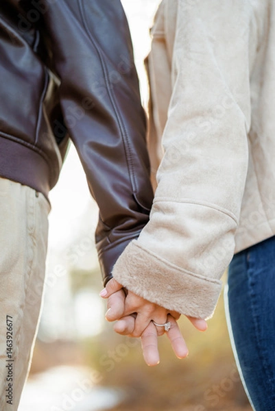 Obraz Couple holding hands together while walking outside. Woman wearing engagement ring crossing fingers with man. Couple's hands close up as symbol of unity and love. Autumn walk of lovers