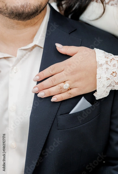 Obraz Bride's hand with wedding ring on finger close-up. Bride put her hand on groom's shoulder, cropped photo. Gold wedding ring with large oval gemstone on bride's finger. Newlyweds in love
