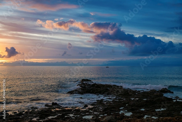 Fototapeta Sunset over rocky coastline with gentle waves and distant sailboat in the serene ocean horizon