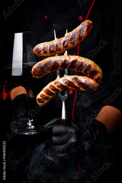 Obraz The chef is holding a metal fork with sausages and a glass of wine. On a black background.