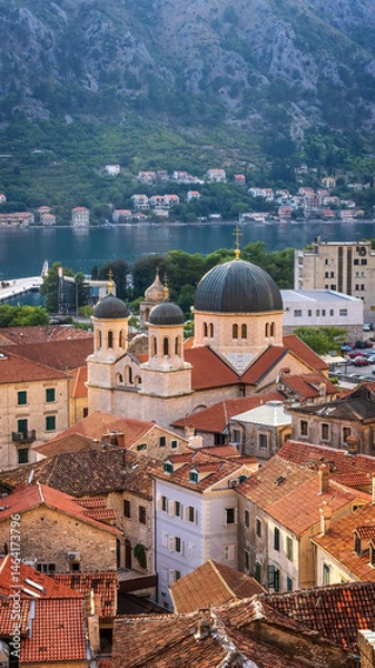 Fototapeta Sunset light bathes the old town of Kotor, Montenegro, highlighting its terracotta rooftops, historic stone buildings, and the iconic dome of St. Nicholas Church near the serene bay.


