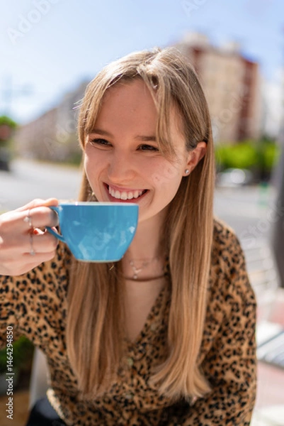 Fototapeta Happy young woman smiling and drinking coffee or tea from a blue cup, sitting at a table in a bar or restaurant in the city during a sunny day