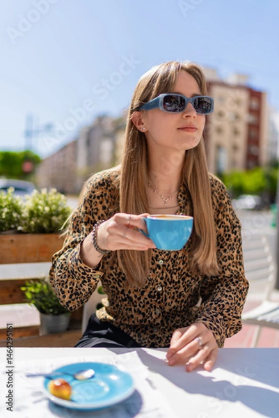 Fototapeta Young woman wearing stylish sunglasses, sipping coffee at a table in an outdoor cafe, soaking up the sun on a beautiful day while enjoying a moment of relaxation and leisure