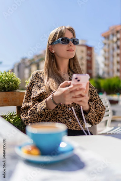 Fototapeta Young woman wearing sunglasses and holding smartphone while sitting at a table with a cup of coffee and a pastry, enjoying breakfast at an outdoor cafe