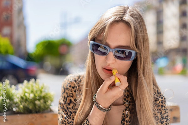 Fototapeta Young blonde woman wearing stylish sunglasses and a trendy leopard print shirt sits at a bar table, applying lip balm while enjoying a sunny day outdoors in the urban setting