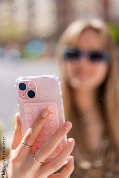 Fototapeta Close up of hands holding a smartphone with a pop it accessory and a transparent case, a young woman with sunglasses is blurred in the background, taking a selfie or recording a video
