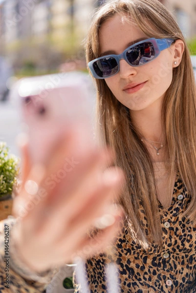 Fototapeta Young woman wearing stylish sunglasses and a vibrant patterned shirt, sitting on a bar terrace, enjoying a sunny day while taking a cheerful selfie with her smartphone