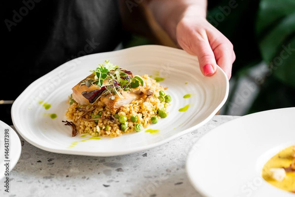 Fototapeta A hand holding a white plate with a gourmet dish featuring a perfectly cooked fish fillet atop grains and green peas, garnished with microgreens and drizzled with a vibrant green sauce
