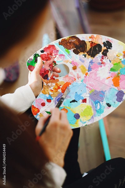 Obraz Top view of female hands holding plastic palette with mixed colors and paint brush. Woman artist in casual clothes in art studio. Creative activity or hobby