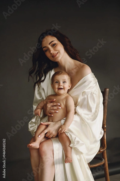 Obraz Loving mother siiting on the chair and hugging her happy baby girl while posing in the studio on black background