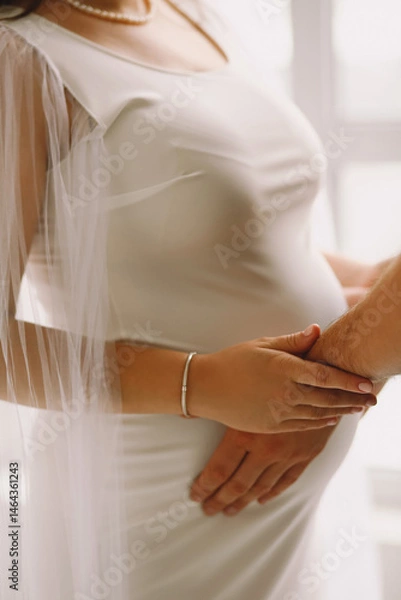 Obraz Close up photo of pregnant woman in white stylish dress and her husband holding their hands on pregnant belly. Family concept