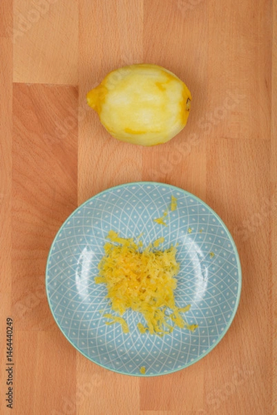 Fototapeta A peeled lemon sits above a light blue decorative plate containing freshly grated yellow lemon zest, all on a light brown wooden surface