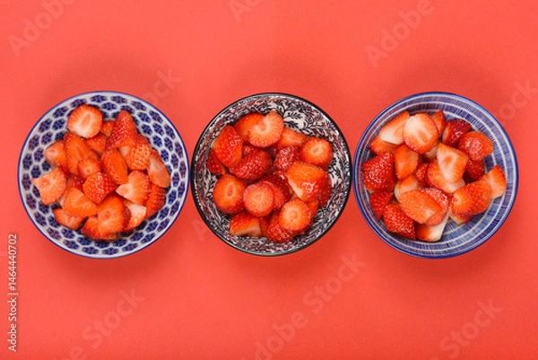 Fototapeta Three small, patterned bowls filled with sliced fresh strawberries are arranged in a row on a vibrant red surface
