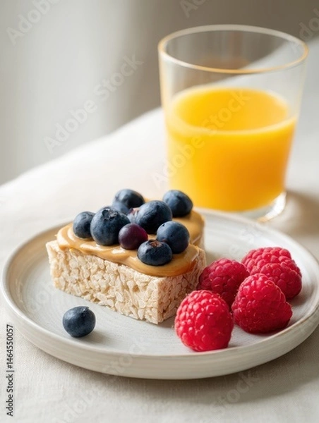 Fototapeta Delicious breakfast with rice cake topped with peanut butter, fresh blueberries, and raspberries alongside a glass of orange juice on a light table.