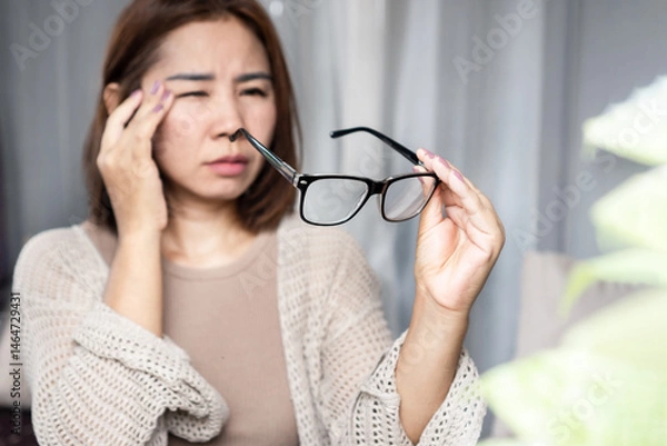 Obraz Asian woman feeling a headache, dizziness, and blurry vision while holding her eyeglasses, showing signs of eye fatigue and vision problems such as nearsightedness or farsightedness
