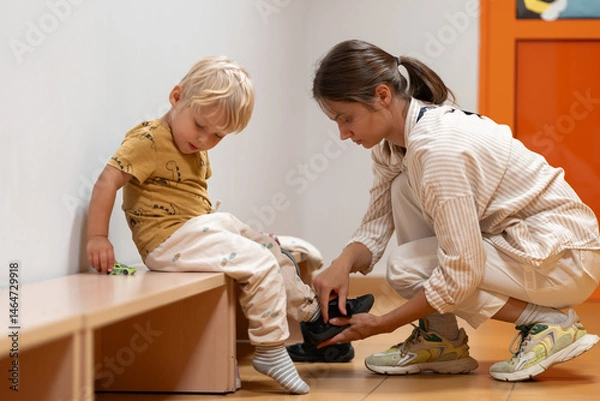 Fototapeta A mother assists her young child with putting on shoes as the child sits on a bench in a kindergarten or daycare hallway. Focus on daily routines, parental care, and getting ready.