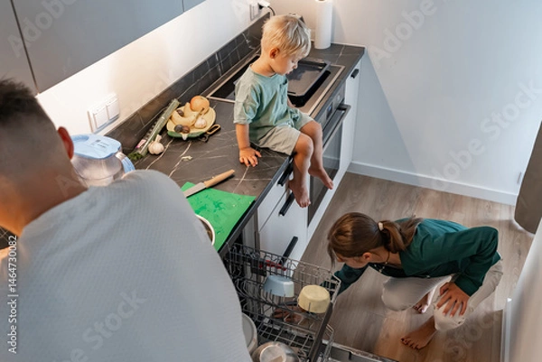 Fototapeta A family scene in a modern kitchen. A woman loads the dishwasher, a man assists, and a young child sits on the counter observing. Depicts daily life, teamwork, and household chores at home.