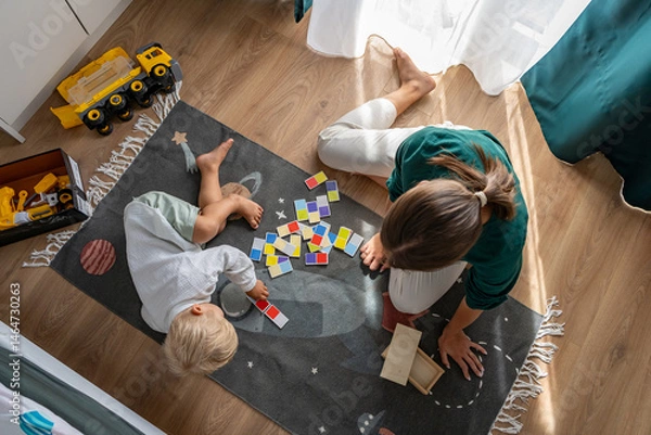 Fototapeta Aerial view of a woman and her young child playing an educational board game with colorful tiles on a space-themed rug. Focus on interactive learning, family bonding, and quality playtime at home.