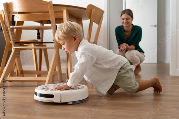 Fototapeta A young blonde child curiously interacts with a robotic vacuum cleaner on the wooden floor at home. The mother observes with a smile in the background, a modern family moment.