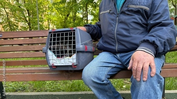 Obraz An elderly retired man holds a cat in a carrier. Pet transportation, traveling with a pet. A close friend of an elderly man