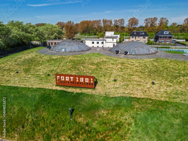 Fototapeta Aerial view of a historic fort with round domes, green grass-covered walls, walking paths, and adjacent buildings, surrounded by trees and open parkland under bright daylight.
