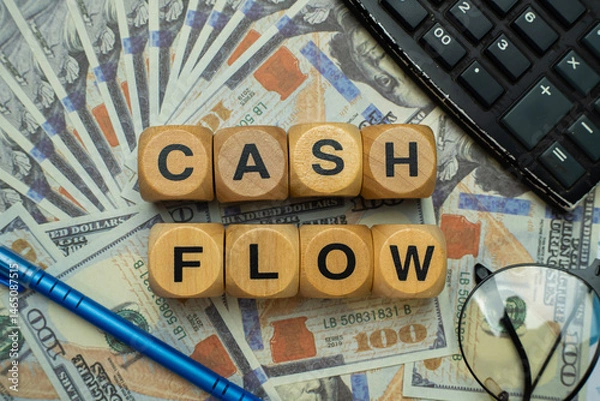 Obraz Wooden block with "CASHFLOW" written on it arranged on top of several $100 bills, surrounded by a pair of glasses, a black calculator and a blue pen. Business and finance concept