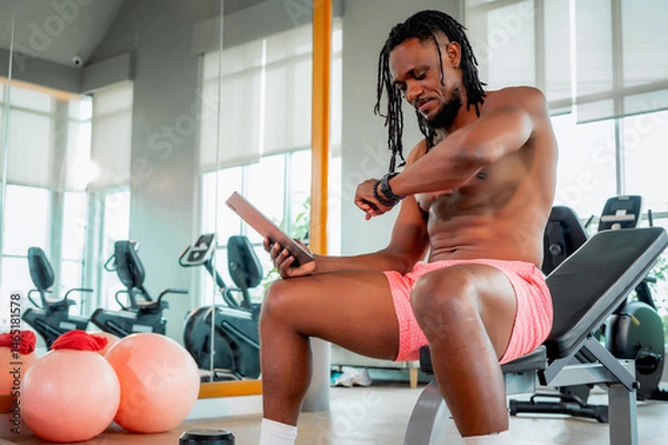 Fototapeta Muscular man takes break at the gym, sitting on bench while checking his smartwatch and holding a tablet. Combining strength training with guided digital instruction. with dumbbells in foreground.