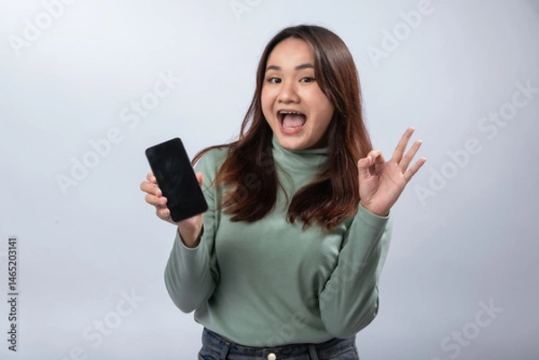 Fototapeta Smiling Asian woman in green sweater holding smartphone with blank screen and making OK hand gesture, winking playfully against plain gray background. Confident approval and tech lifestyle concept