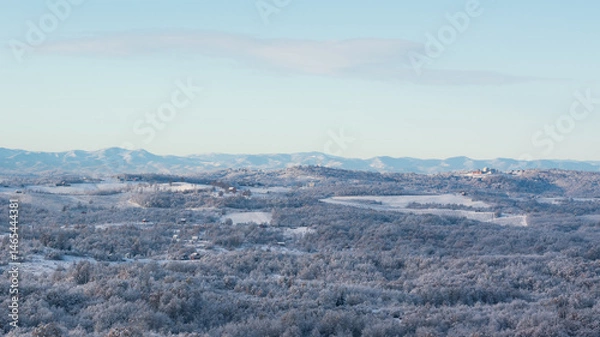 Fototapeta Hilly village covered with frost after cold winter night, landscape with deciduous forest and fields, mountain range in distance