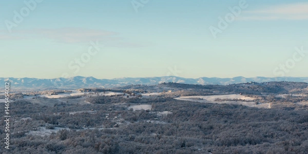Fototapeta Hilly village covered with frost after cold winter night, rural landscape with houses, deciduous forest and fields, mountain range in distance