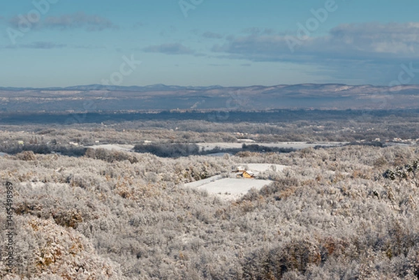 Fototapeta Wintry landscape of village surrounded with forest and distant hills in haze