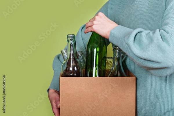Fototapeta Hand placing green glass bottle into box filled with used bottles for sorting and recycling at home