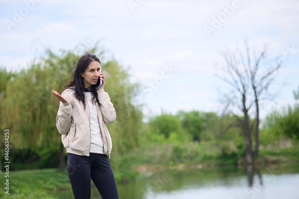 Fototapeta Smiling woman making a video call in park and pointing upward