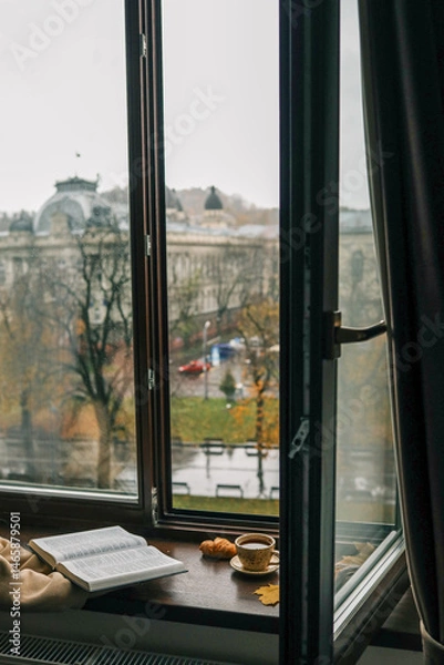 Obraz Person sitting by a window reading a Bible with a cup of coffee and a croissant on the windowsill, overlooking a rainy autumn park