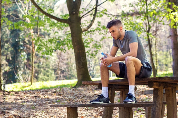 Fototapeta Young man sitting on bench at the park and relaxing after jogging