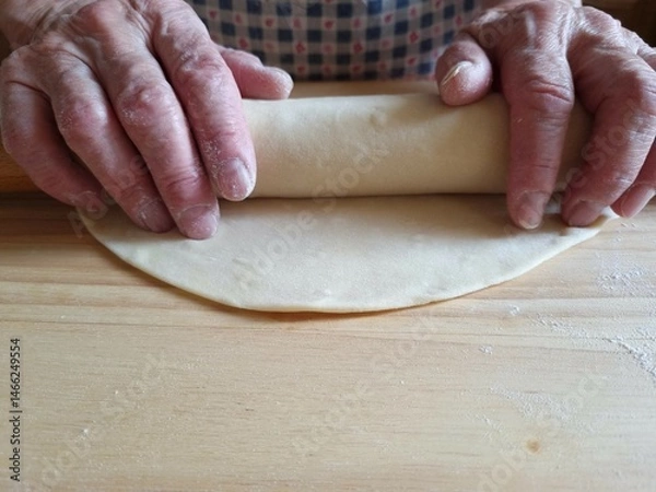 Obraz An elderly lady rolls out dough with a wooden rolling pin on a table. Close-up of the elderly lady's hands. Home cooking