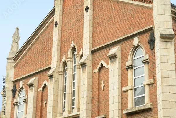 Fototapeta Neo Gothic Revival church facade with red brick walls and light stone trim featuring pointed arch lancet windows decorative buttresses and tracery under an overcast sky