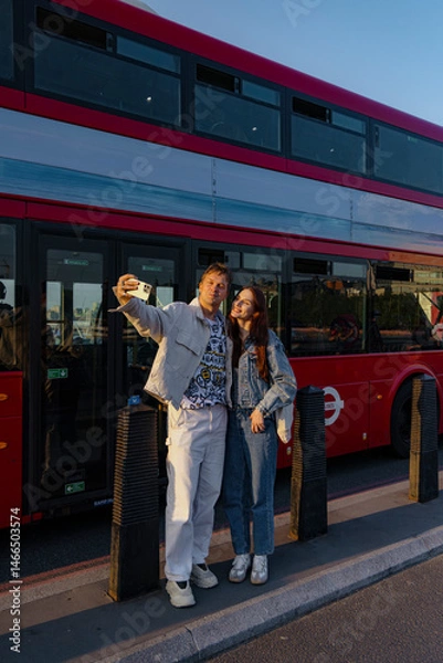 Obraz A young couple fills the air with laughter and joy as they pose together for a cheerful selfie in front of a classic London bus, capturing a delightful moment of happiness amidst the vibrant city