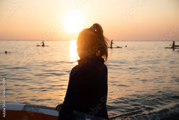 Fototapeta An elderly woman rides paddleboarding in the ocean. Senior female is waiting for a wave on a surfboard. Active old age. Old girl is sitting on a board in the sea at sunset, dressed in a swimming suit