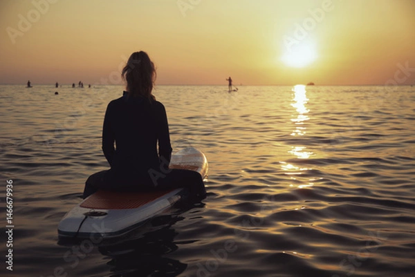 Fototapeta An elderly woman rides paddleboarding in the ocean. Senior female is waiting for a wave on a surfboard. Active old age. Old girl is sitting on a board in the sea at sunset, dressed in a swimming suit